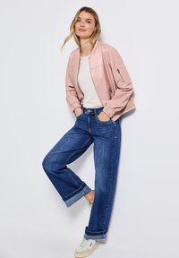 Veste bombardier rose clair avec des poignets élastiques, associée à un jean large bleu avec des ourlets roulés, et des baskets blanches avec des accents beiges.