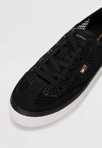 Baskets en mesh noir avec lacets noirs, semelle blanche et petit logo rectangulaire rouge, blanc et or sur le côté.