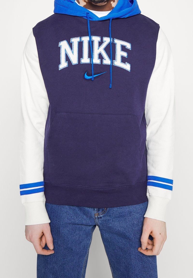 Homme portant un sweat à capuche Nike bleu marine et bleu avec des manches blanches et des poignets rayés bleus, associé à un jean bleu, mains détendues sur les côtés.