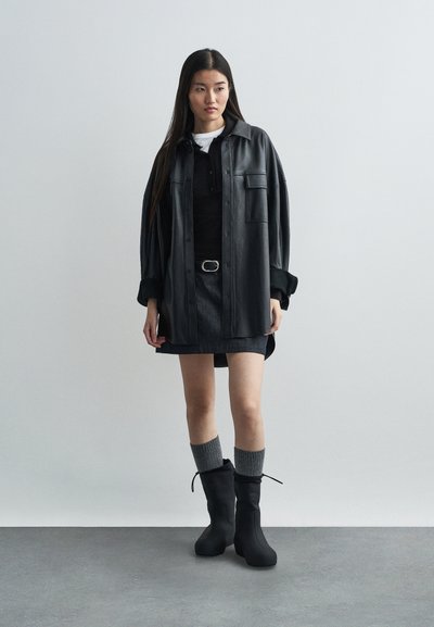 Cămășă din piele neagră supradimensionată, top negru, fustă scurtă, șosete tricotate gri și bocanci negri cu finisaj texturat și detaliu cu șnur.