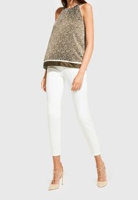 Haut beige sans manches avec un motif à pois et une bordure foncée, associé à un pantalon blanc ajusté et des chaussures à talons hauts couleur chair.