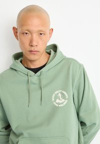 Sweatshirt verde menta feito de um tecido macio, com um bolso frontal e um logótipo circular em cor creme com texto. Detalhe do capuz com cordão visível.
