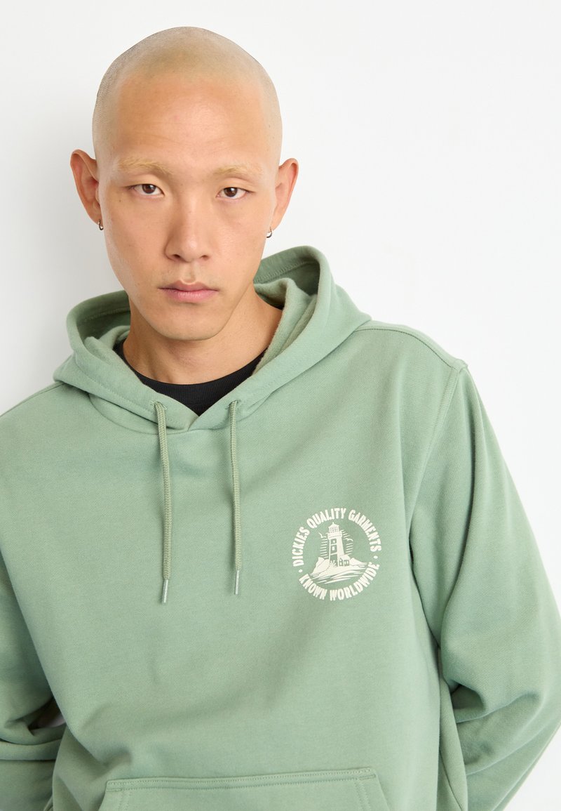 Mint green hoodie made from soft fabric, featuring a front pocket and a circular logo in cream color with text. Drawstring hood detail visible.