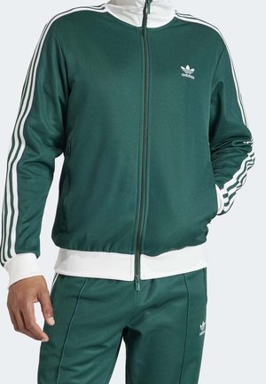 Mann trägt dunkelgrünen Adidas-Trainingsanzug mit weißen Streifen an Ärmeln und Hose, weißen Bündchen und Reißverschluss vorne an der Jacke.
