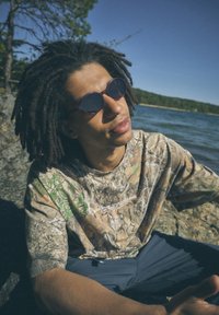 Μπλούζα με παραλλαγή σε πράσινα και καφέ σχέδια, στρογγυλά μπορντό γυαλιά ηλίου, κάθεται δίπλα σε βραχώδη όχθη λίμνης κάτω από καθαρό γαλάζιο ουρανό.