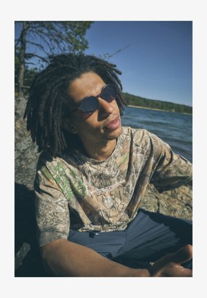 Majica s kamuflažno potiskom v zelenih in rjavih vzorcih, okrogle bordo rdeče sončna očala, sedi ob skalnati obali jezera pod jasnim modrim nebom.