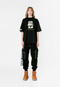 Camiseta negra de gran tamaño con estampado gráfico, combinada con pantalones cargo negros con bolsillos con cremallera y botas tan con cordones.