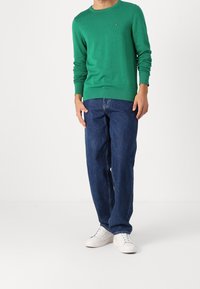 Homme portant un pull vert à manches longues, un jean bleu et des baskets blanches, debout devant un fond blanc uni.