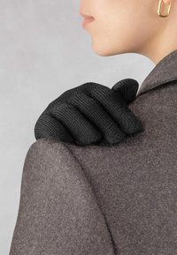 Schwarze Strickhandschuhe liegen auf der Schulter eines grauen Wollmantels und präsentieren ein strukturiertes Design und eine klare Silhouette. Der Hintergrund ist hellgrau.