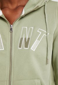 Sudadera con capucha verde claro con cremallera, hecha de tela suave, con letras bordadas en blanco y metalizadas "ANT" en el frente, con capucha ajustable con cordón.