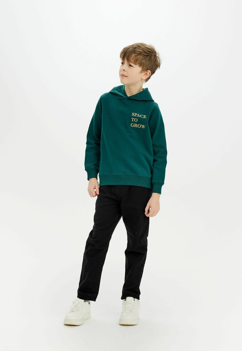 Grön hoodie med texten "SPACE TO GROW" i guld, kombinerat med svarta byxor och vita sneakers. Tyget verkar mjukt och avslappnat.