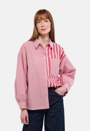 Femme portant une chemise ample à boutons rouge et blanche à rayures verticales mélangées, avec un pantalon bleu foncé, posant avec une main dans la poche.