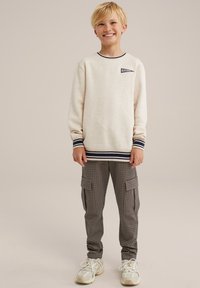 Vaaleahiuksinen poika hymyilee, päällään vaaleanruskea collegepaita, jossa on tummansinisiä raitoja ja "Blue Ridge" -logo, ruudulliset cargo-housut ja valkoiset tennarit, seisten yksinkertaista taustaa vasten.