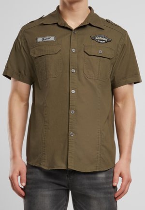 Mand iført en kortærmet olivengrøn skjorte med knapper, to brystlommer og broderede mærker på den øverste del af brystet.