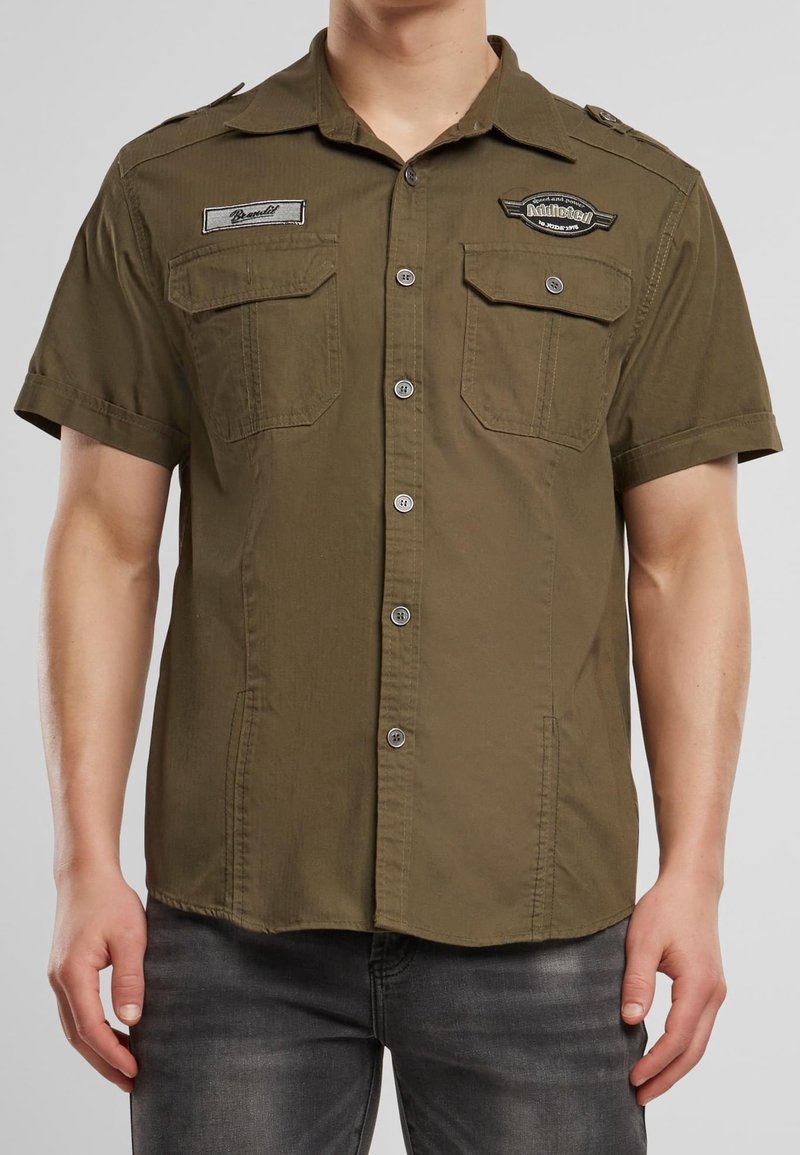 Homme portant une chemise boutonnée à manches courtes de couleur vert olive avec deux poches sur la poitrine et des écussons brodés sur la partie supérieure de la poitrine.