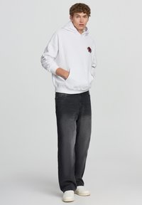 Felpa bianca con una rosa rossa ricamata sul petto, abbinata a jeans neri e sneakers bianche. Vestibilità comoda con tasca a marsupio.