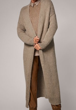 Personne portant un long cardigan beige en maille épaisse sur un pull marron, une chemise beige à col, et un pantalon marron, debout devant un fond uni.