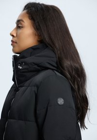 Sort pufferjakke med høj krave, lynlåslukning og detalje med sidelynlås. Blød tekstur med diskret branding på ærmet.