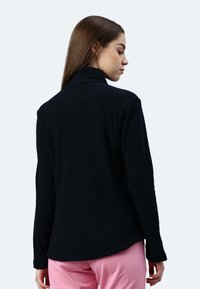 Giacca in pile nera con colletto alto e maniche lunghe, realizzata in un materiale morbido e testurizzato e con una vestibilità dritta, abbinata a pantaloni rosa.