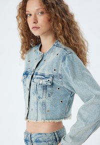 Kappad jeansjacka i ljusblått med silvriga öglor och franskant. Har två bröstfickor och knappstängning.