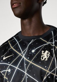 Juodas sportinis marškinėliai su kosmine dizaino, kuriame yra baltos ir auksinės linijos bei taškai, ir išsiuvinėtas liūto logotipas ant krūtinės. Taip pat matomas Nike prekės ženklas.
