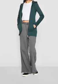 Felpa blu verde con zip e tasche frontali, abbinata a pantaloni grigi larghi e sneakers nere. Design semplice e rilassato con un top bianco corto.