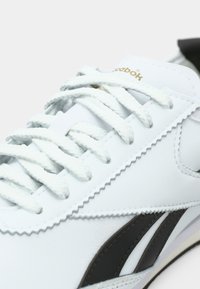 Sneaker din piele albă cu dungi laterale negre, șireturi albe și logo auriu pe limbă, prezentat în detaliu apropiat.