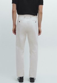 Lichtbeige broeken met een gladde textuur, voorzien van twee achterzakken en bruine knoopaccenten, gecombineerd met een zwart shirt en zwarte schoenen.