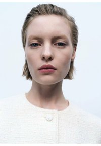 Weiß strukturierte Jacke mit rundem Ausschnitt und Knopfverschluss, getragen von einem Modell mit kurzem Haar und natürlichem Make-up vor einem hellen Hintergrund.