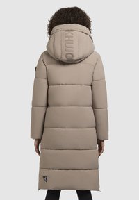 Bežs pūkains mētelis ar garu siluetu, raksturīgs ar horizontālām quiltējuma daļām, lielu kapuci un logotipu aizmugurē.