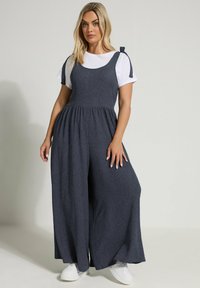Marinblå ribbad jumpsuit med vida ben, justerbara knytband och en åtsittande bodice, kombinerad med en vit T-shirt och vita sneakers.