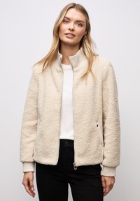 Frau trägt eine cremefarbene Fleecejacke mit Reißverschluss über einem weißen Hemd und schwarzen Hosen, steht vor einem einfarbigen grauen Hintergrund.