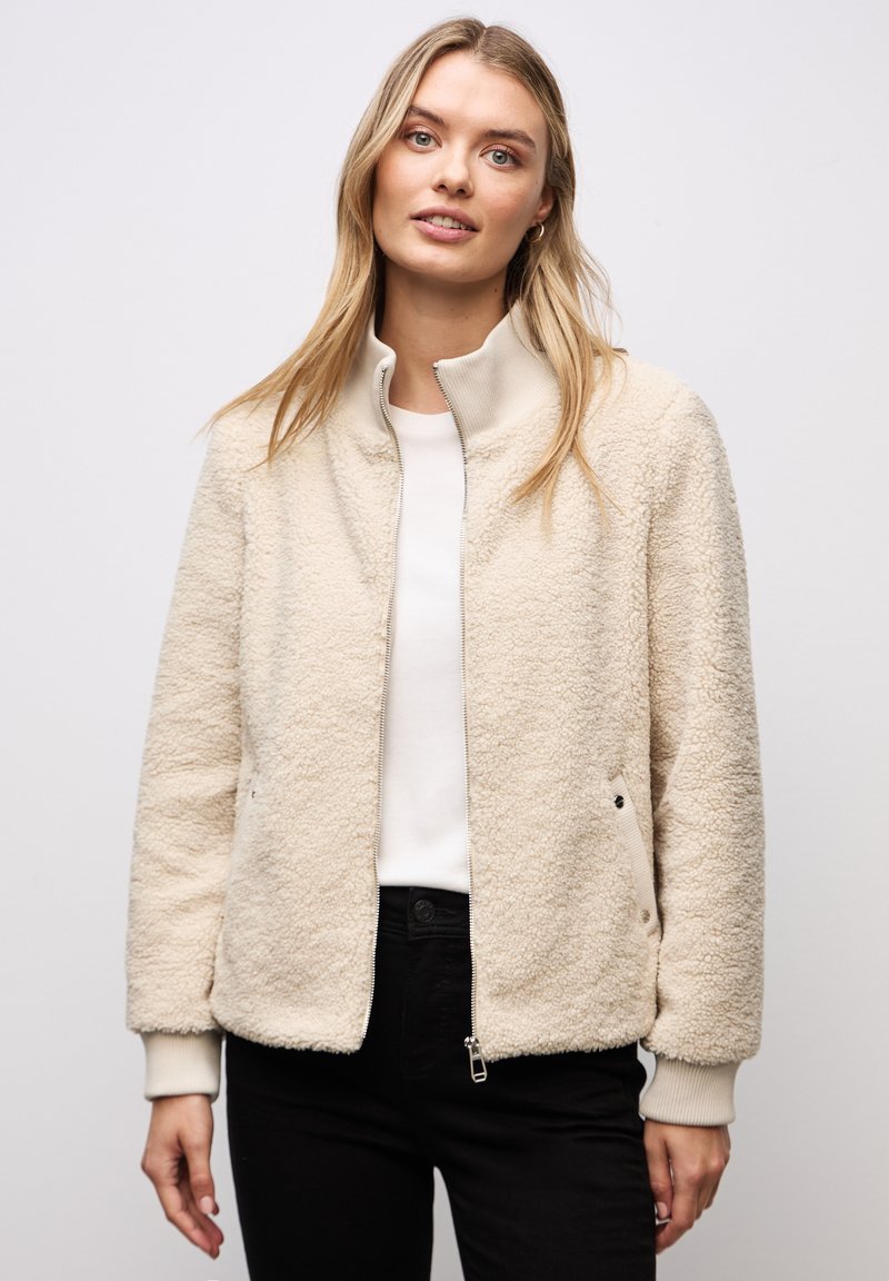 Frau trägt eine cremefarbene Fleecejacke mit Reißverschluss über einem weißen Hemd und schwarzen Hosen, steht vor einem einfarbigen grauen Hintergrund.