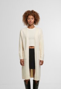 Krémový dlouhý cardigan vyrobený z měkké, texturované látky, s dvěma předními kapsami a otevřeným předním designem, kombinovaný s krátkým topem a černou sukní.