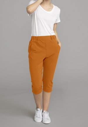 Femme portant un T-shirt blanc à manches courtes, un pantalon court orange avec des poches, et des baskets blanches sur un fond gris uni.