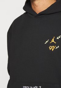 Sudadera negra con llamas naranjas, logo de Jumpman, iniciales "CW" estilizadas en el pecho y texto blanco "¿POR QUÉ NO?" en el bolsillo.