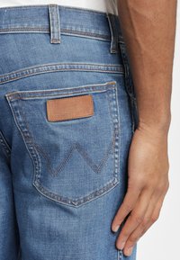 Primer plano de la mano derecha de una persona descansando junto a un bolsillo trasero de unos vaqueros de denim azul con costuras marrones y una etiqueta de cuero.