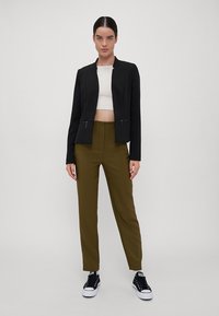 Black fitted jacket with a cropped design, paired with olive green trousers. Model wears a white top and black sneakers.