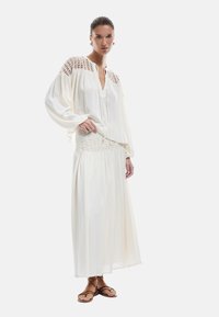 Blouse blanche fluide avec des accents en crochet sur les épaules, associée à une jupe maxi blanche à volants. Des sandales à lanières brunes complètent le look.