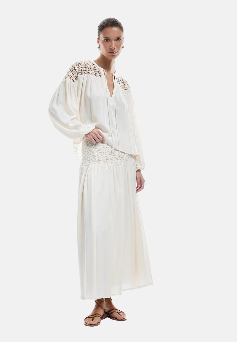 Blouse blanche fluide avec des accents en crochet sur les épaules, associée à une jupe maxi blanche à volants. Des sandales à lanières brunes complètent le look.