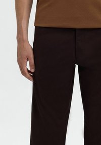 Bruine textiele top in combinatie met donkerbruine broek, met een gladde textuur en een getailleerde pasvorm, met een hand die op de zijkant van de broek rust.