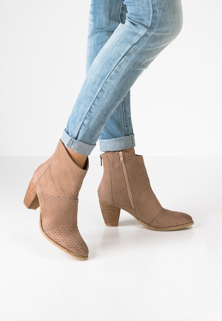 Brown ankle boots with a textured design, pointed toe, and side zipper. Set on a stacked heel, paired with light blue skinny jeans.