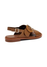 Sandal in pelle marrone con soletta piatta, punta aperta, cinturino alla caviglia regolabile e una grande fibbia rotonda come dettaglio laterale.