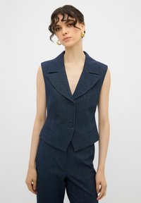 Ärmlös marinblå denimblazer med stor sjalkrage, enknappsstängning och två framfickor. Texturerad tyg med skräddarsydd passform.