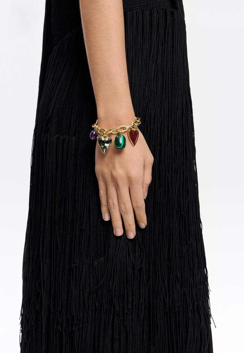 Pulsera de cadena de oro con cuatro encantos en forma de corazón de colores: transparente, verde, rojo y púrpura, contra una prenda negra con flecos.