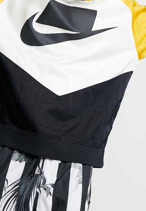 Hvid og sort Nike sweatshirt med gule ærmer over sort-hvide bukser med blomstermønster.