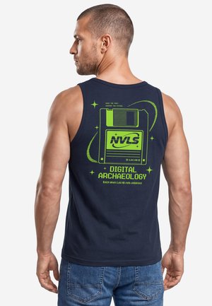Man wearing navy tank top with bright green floppy disk graphic and text "Digital Archaeology" on the back, facing away and looking left.