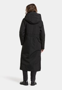 Lange zwarte pufferjas met een capuchon, een Getailleerde taille en zijsplitsen, gemaakt van een gladde, duurzame stof. Gekozen in combinatie met donkere broeken en zwarte laarzen.