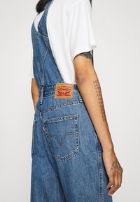 Blå denimoverall med justerbara axelremmar, en Levi's etikett på baksidan och en klassisk fickdesign. Tyget verkar något slitet.