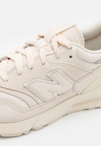 Beige lädersneaker med nätaccenter, texturerad yta och en framträdande "N"-logotyp. Har platta skosnören och gummisula för stöd.